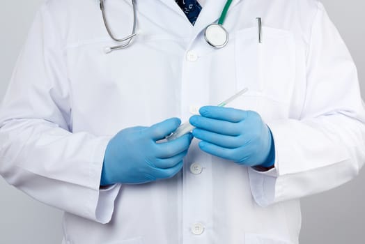 male doctor in a white coat and tie stands and holds a plastic syringe with a needle on a white background, wearing blue sterile medical gloves in his hands the concept of vaccination against diseases