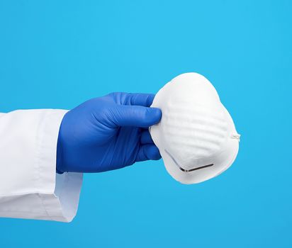 doctor in a white coat, blue latex sterile gloves holds textile disposable  masks in his hand on a blue background, additional protection against viruses during an epidemic, pandemic