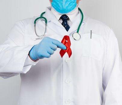 doctor in white uniform and sterile latex gloves holds a red ribbon-symbol of the fight against disease AIDS and vasculitis, close up