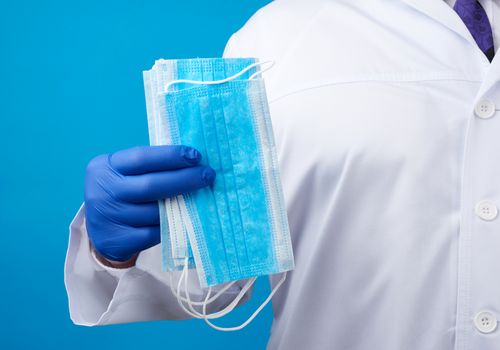 doctor in a white coat, blue latex sterile gloves holds textile disposable medical masks in his hand on a blue background