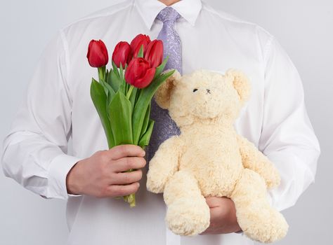 adult man in a white shirt holds a bouquet of red blooming tulips and a white teddy bear, white background. Happy birthday, date, greetings concept