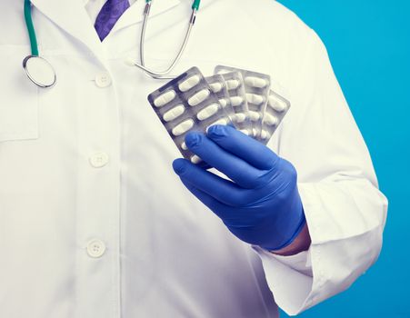 male hand with blue sterile gloves holds a stack of pills in a blister, concept for drug treatment of diseases, blue background