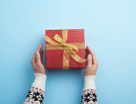 female hands holds a red box tied with a golden ribbon, concept of congratulations and gifts, top view, blue background