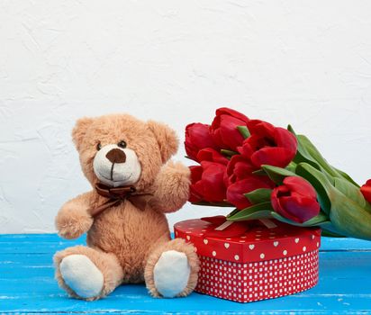 cute brown teddy bear sits on a blue wooden background, bouquet of red tulips, red box, festive backdrop for birthday, Valentine's day, anniversary