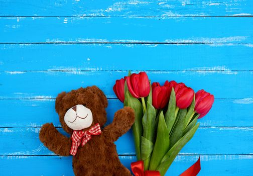cute brown teddy bear and bouquet of red blooming tulips with green stems and leaves tied with a red silk ribbon, flowers lie on a blue wooden background, top view, copy space