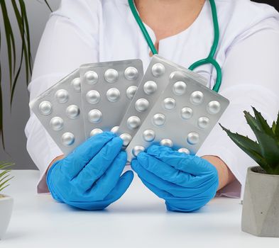 female hand with blue sterile gloves holds a stack of pills in a blister, concept for drug treatment of diseases, doctor sits in an armchair at a desk, office