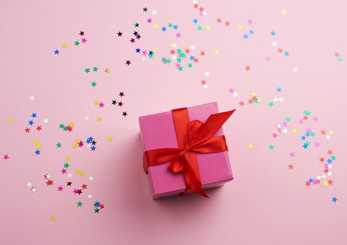 beautiful pink closed box with a big bow on a pink background with multi-colored sparkles, concept of congratulations and surprise, top view