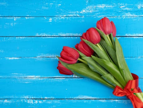 bouquet of red blooming tulips with green stems and leaves tied with a red silk ribbon, flowers lie on a blue wooden background, top view