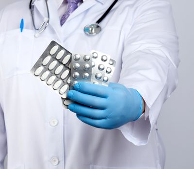 doctor therapist is dressed in a white uniform coat and blue sterile gloves is standing and holding a stack of pills in blister packs, white background