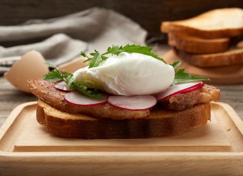 sandwich on toasted white slice of bread with poached eggs, green leaves of arugula and radish, morning breakfast on a brown wooden board