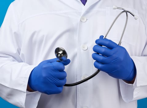 adult male doctor in a white medical coat stands and holds a black stethoscope on a blue background, close up