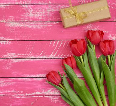 bouquet of red blooming tulips with green leaves, wrapped gift in brown craft paper and tied with silk ribbon on a pink wooden background, top view. Festive backdrop for birthday, Valentine's day