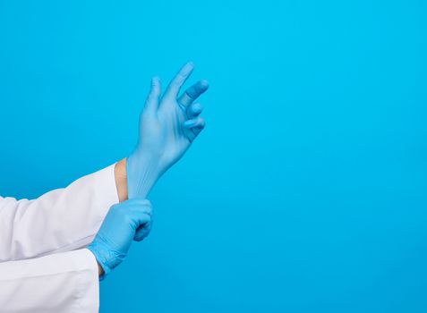 female doctor in a white coat puts on her hands blue latex sterile medical gloves on her hands, blue background, place for text