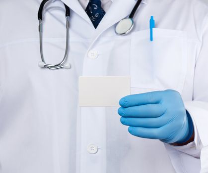 doctor therapist is dressed in a white robe uniform and blue sterile gloves is standing and holding a stack of empty white paper business cards, place for inscription