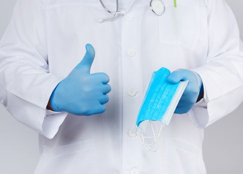 doctor in a white coat, blue latex sterile gloves holds textile medical masks in his hand on a white background, other hand shows like, protective accessory against viruses and bacteria