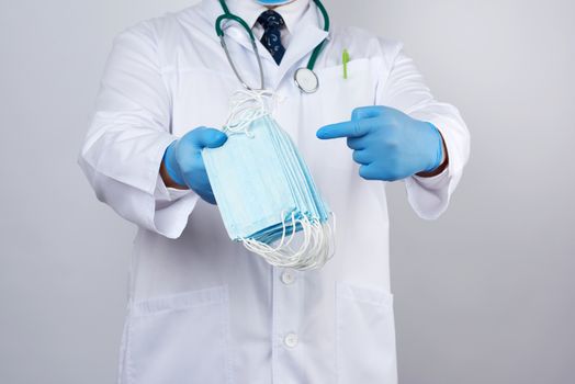 doctor in a white coat, blue latex sterile gloves holds textile medical masks in his hand on a white background, other hand shows like, protective accessory against viruses and bacteria