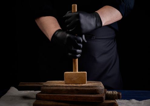 male cook in black uniform holds a wooden hammer for beating meat on a vintage cutting board, black background