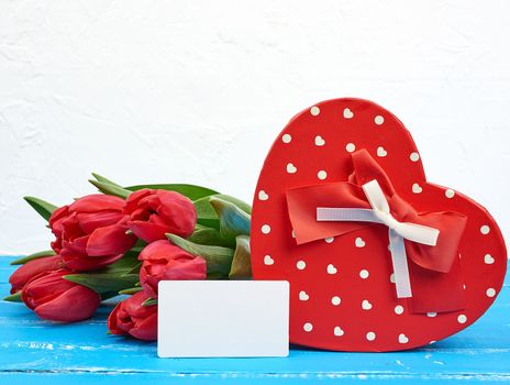 empty rectangular white paper business card, bouquet of red tulips and a heart-shaped cardboard box on a blue wooden background. Festive backdrop for birthday, Valentine's day, anniversary