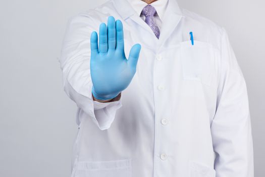 male doctor in a white coat and blue sterile gloves shows a stop gesture with his right hand, white background
