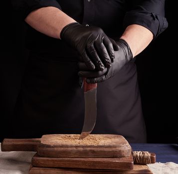 male cook in black uniform and black latex gloves holds a vintage sharp knife over a cutting board, black background