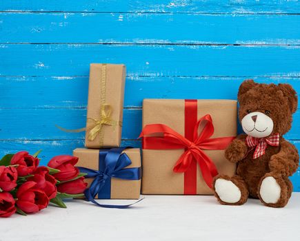 cute brown teddy bear sits on a blue wooden background, bouquet of red tulips, red box, festive backdrop for birthday, Valentine's day, anniversary