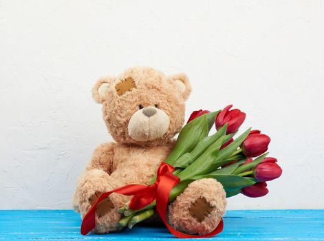 cute brown teddy bear with patches sits on a blue wooden background and holds in his paw a bouquet of red tulips, festive backdrop for birthday, Valentine's day, anniversary