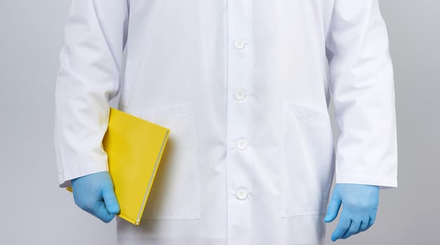 male doctor in a white coat with buttons, wearing blue sterile gloves, holding a yellow paper notebook in his hand on a white studio background