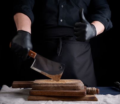 male cook in black uniform and black latex gloves holds a large sharp meat knife over a cutting board, another hand shows like gesture, black background