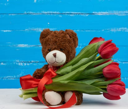 cute brown teddy bear with patches sits on a blue wooden background and holds in his paw a bouquet of red tulips, festive backdrop for birthday, Valentine's day, anniversary