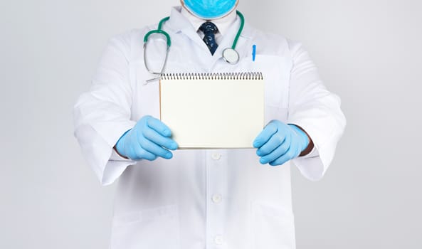 male doctor in a white coat and medical blue gloves holds a notebook with blank white sheets on a white background, place for an inscription