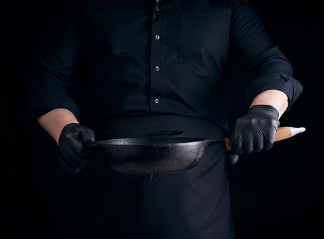 male cook in black uniform and latex gloves holds an empty round vintage black cast iron pan in front of him, low key