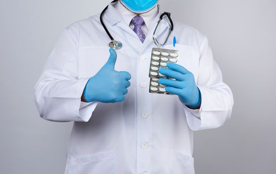 doctor therapist is dressed in a white uniform coat and blue sterile gloves is standing and holding a stack of pills in blister packs, white background