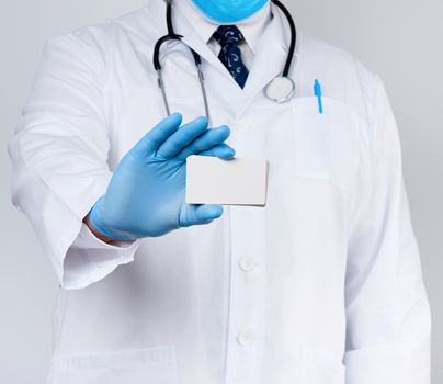 adult doctor therapist is dressed in a white robe uniform and blue sterile gloves is standing and holding a stack of empty white paper business cards, place for inscription