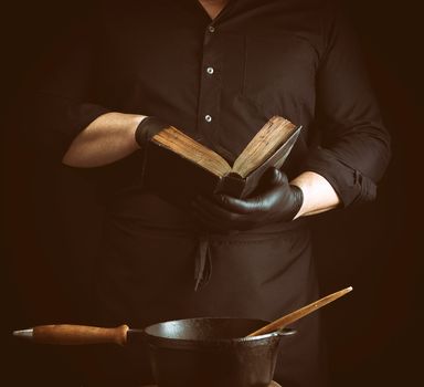 male cook in a black uniform holds an old cookbook with recipes, on a table is a cast-iron frying pan with a wooden handle, low key
