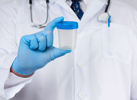 male doctor in a white coat and tie stands and holds a plastic container for urine specimen, wearing blue sterile medical gloves