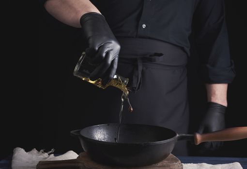 chef in black latex gloves pours olive oil from a transparent bottle into a black cast-iron frying pan, close up