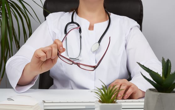 woman doctor in a white uniform, disposable medical mask sits in a chair at the table and works, holds glasses in his hands, office