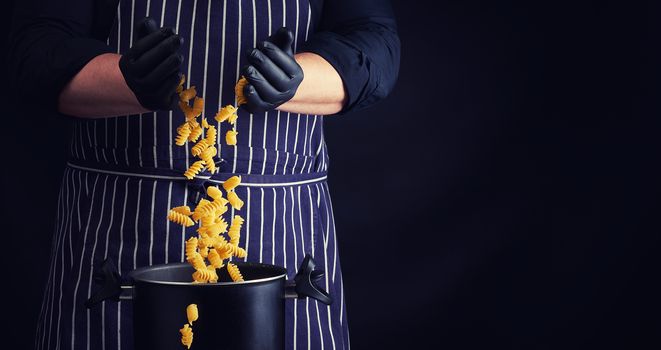 chef in black latex gloves, striped apron pours raw fusilli pasta into a metal pan, cooking process, twisted pasta frozen in the air, copy space