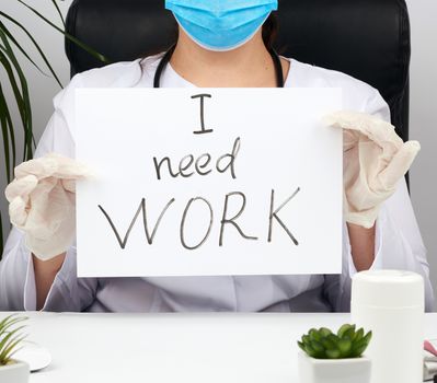 woman doctor in a white coat, sterile medical gloves holds a poster with the inscription I need work, concept of unemployment and job search