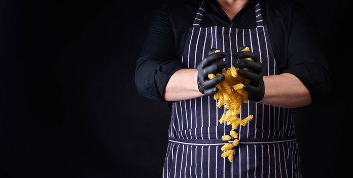 male cook in a striped apron and black latex gloves throws raw fusili pasta down, dark background, place for napsi, the product froze in the air