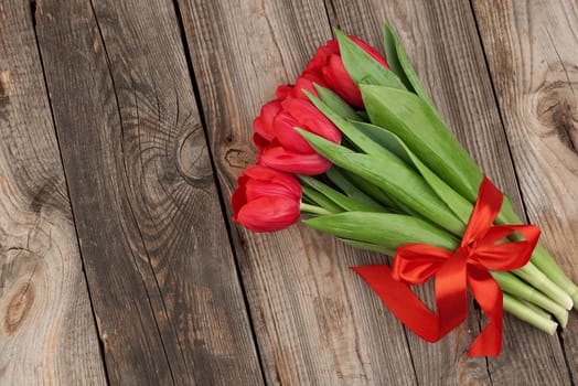 bouquet of red blooming tulips with green stems and leaves tied with a red silk ribbon, flowers lie on a gray wooden background, top view