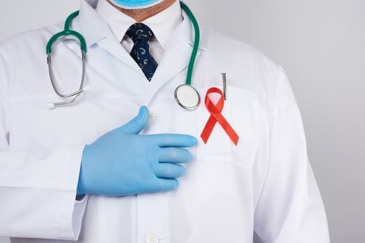 male doctor in a white coat and tie is standing on a white background, red silk ribbon is hanging on his chest, and blue sterile medical gloves are on his hands, symbol of the fight against disease AIDS and vasculitis
