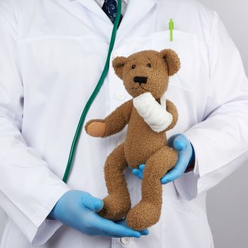 pediatrician in a white coat, blue latex gloves holds a brown teddy bear with a bandaged paw, toy treatment