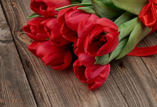 bouquet of red blooming tulips with green stems and leaves tied with a red silk ribbon, flowers lie on a gray wooden background, close up