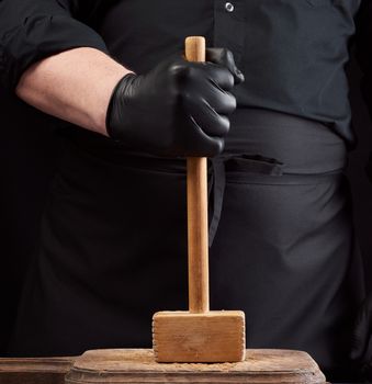 cook in black uniform holds a wooden hammer for beating meat on a vintage cutting board, black background