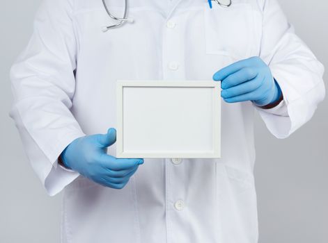 male doctor in a white medical coat and blue latex gloves holds an empty white frame for an inscription, template for a designer