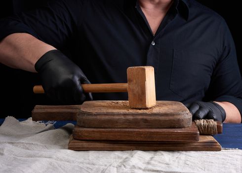 male cook in black uniform holds a wooden hammer for beating meat on a vintage cutting board, black background