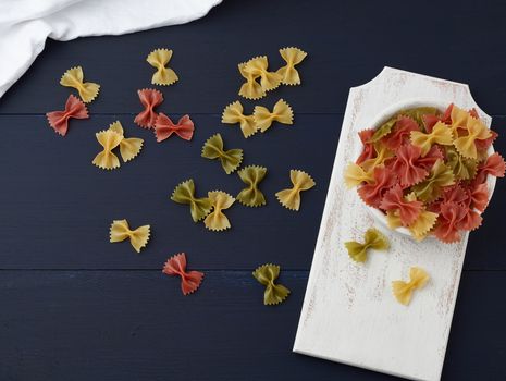 raw pasta in the form of bows in a wooden round plate and white textile napkin on a blue wooden background, top view. Vegetarian nutrition. Italian cuisine