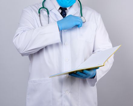 doctor in a white coat with buttons, wearing blue sterile gloves, holding a paper open notebook in his hand on a white studio background