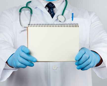 male doctor in a white coat and medical blue gloves holds a notebook with blank white sheets, place for an inscription